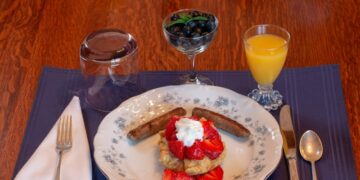 Close up of a breakfast plate with strawberries on top of a pancake, orange juice, blueberries, and fine silverware
